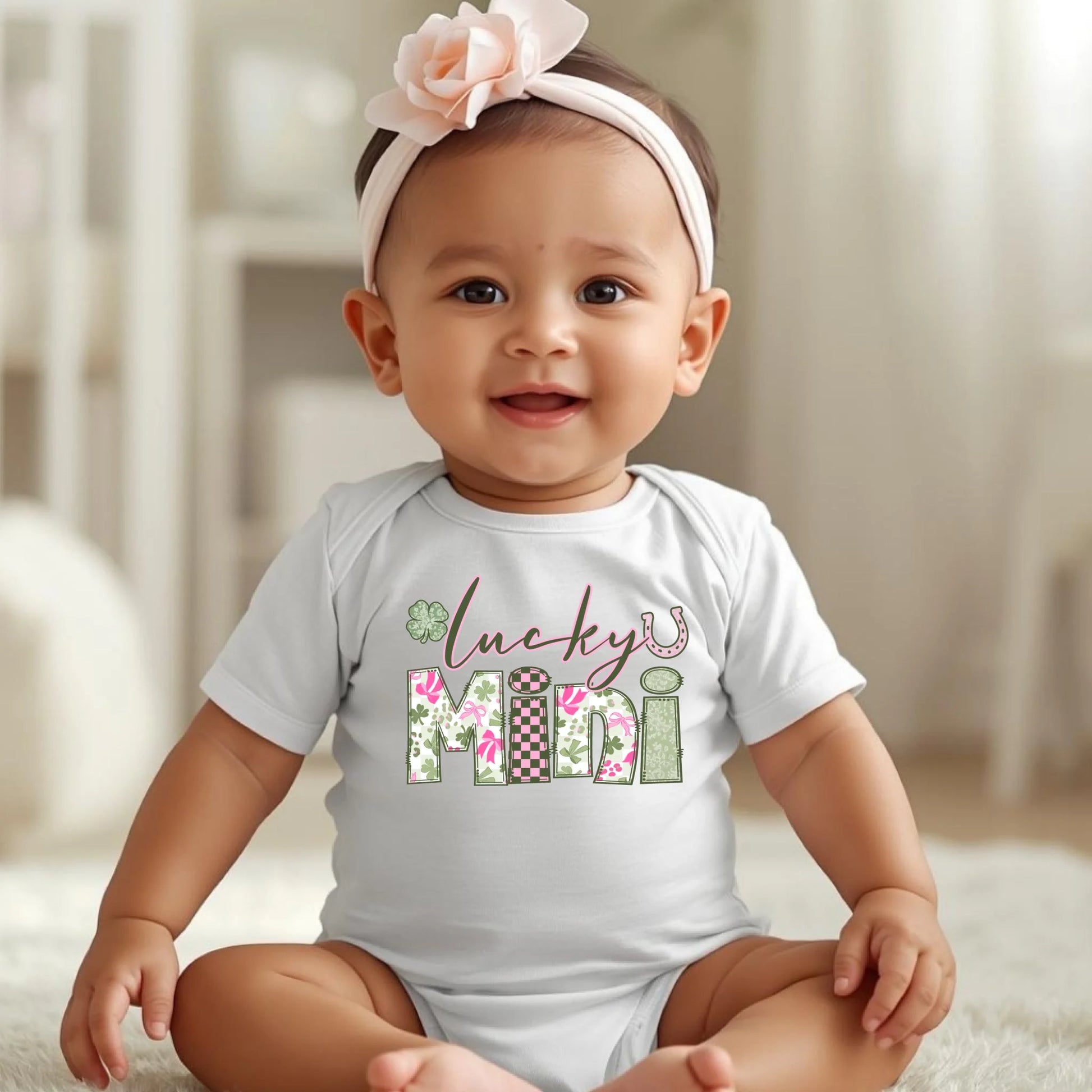 Baby wearing a white onesie with 'lucky mini' print, sitting on a soft surface.