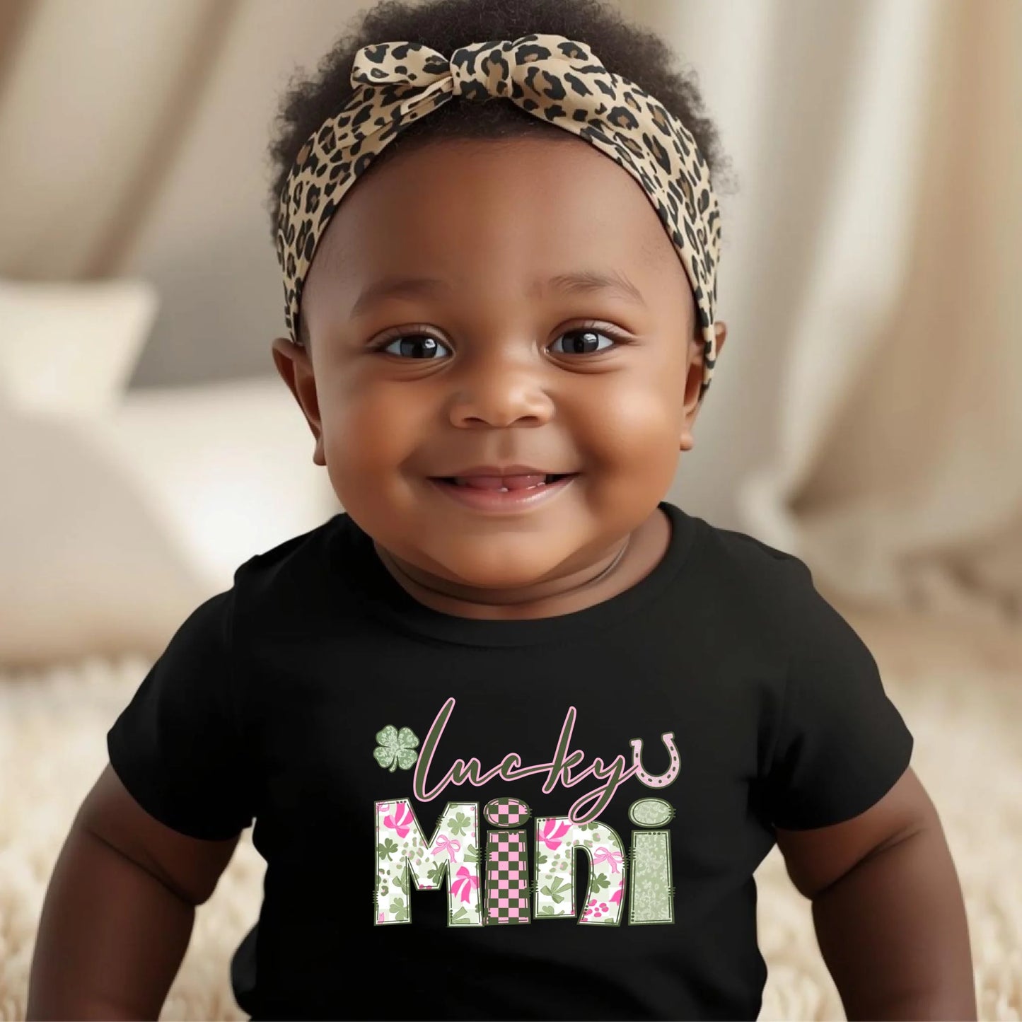 Baby wearing a black shirt with 'Lucky Mini' text and a leopard print headband.