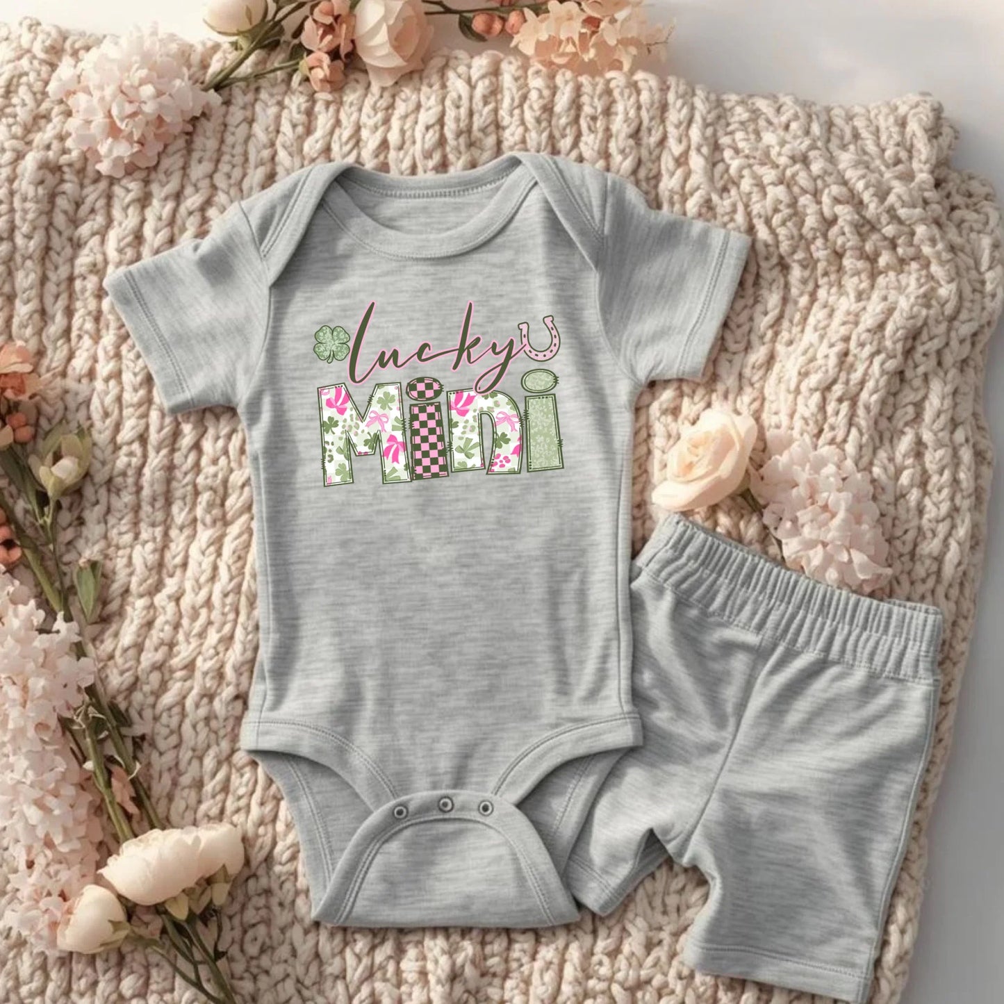 Gray baby onesie with 'Lucky Mini' print on a textured surface with flowers.
