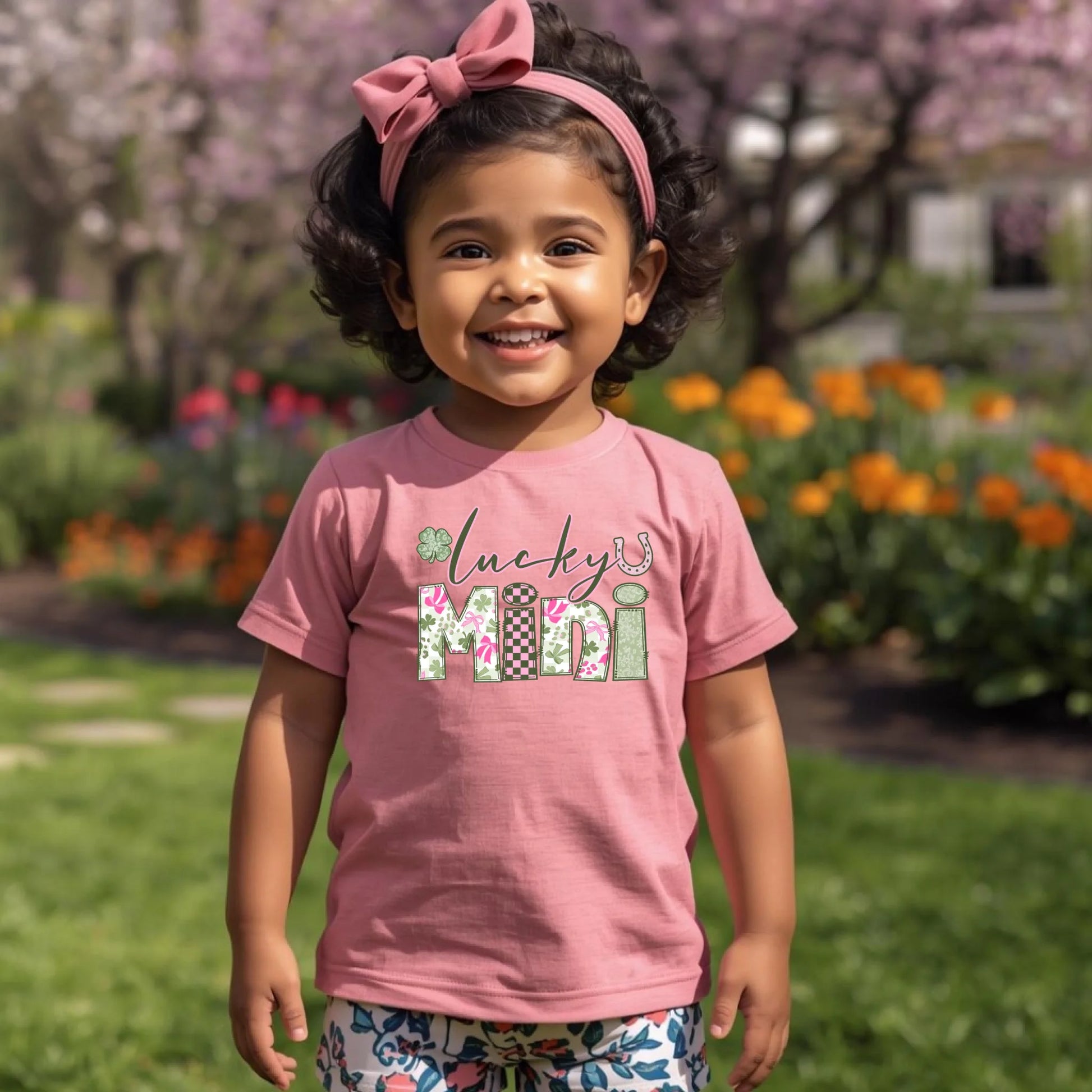 Young girl wearing a pink shirt with 'lucky mini' text, standing in a garden with flowers and trees.