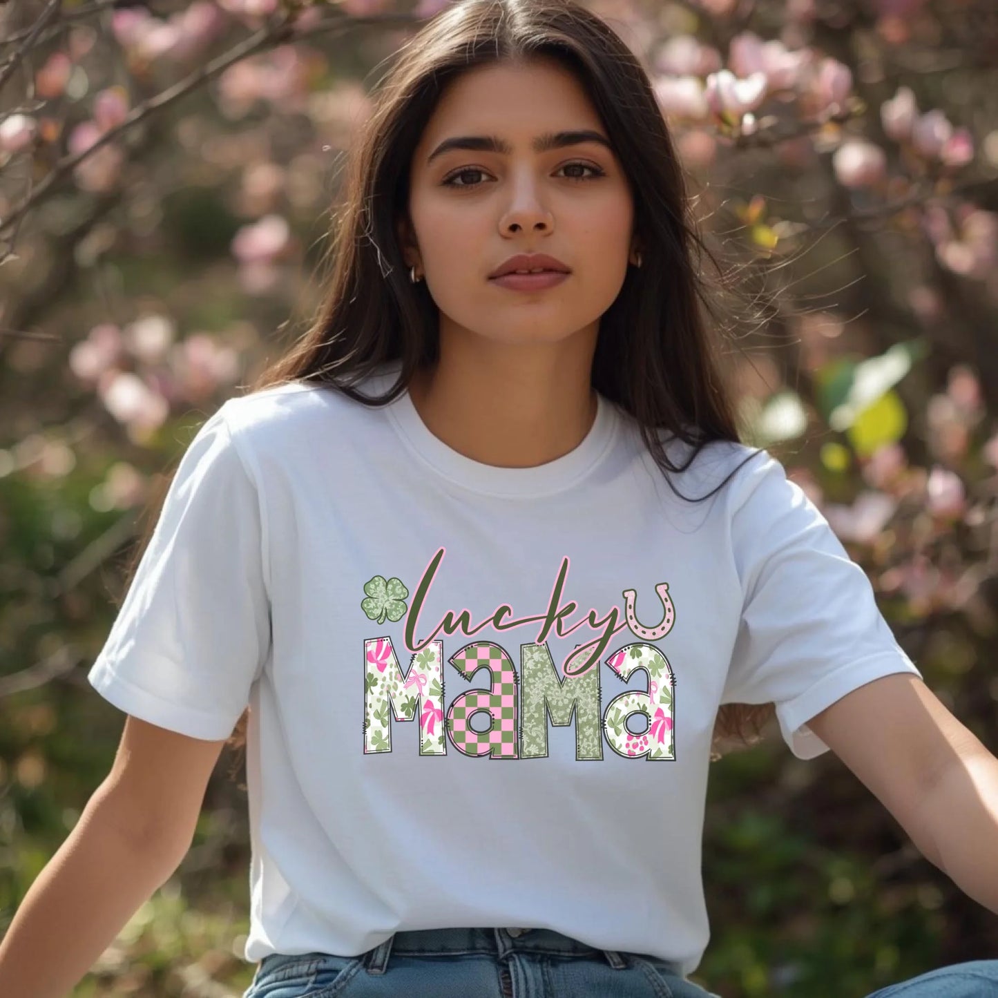 Woman wearing a white t-shirt with 'lucky mama' text in a floral design, standing outdoors with blurred pink flowers in the background.
