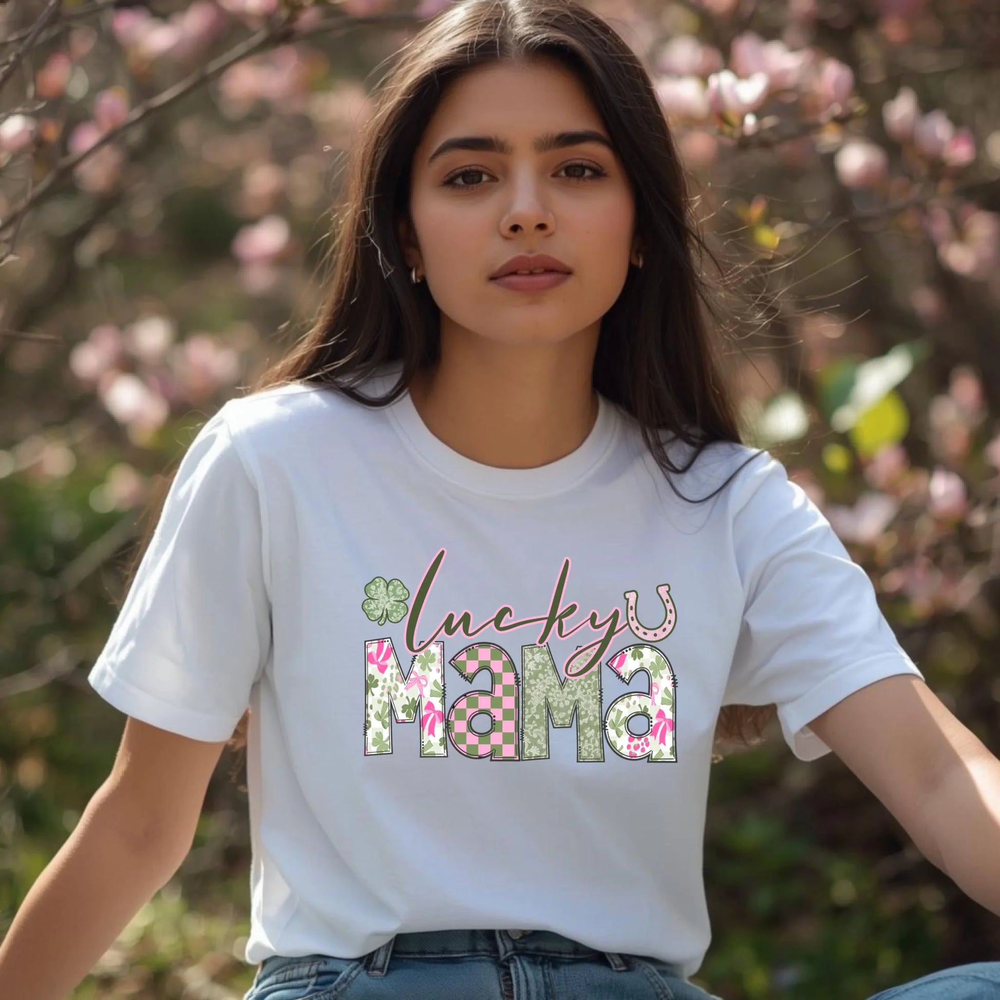 Woman wearing a white t-shirt with 'lucky mama' text in a floral design, standing outdoors with blurred pink flowers in the background.