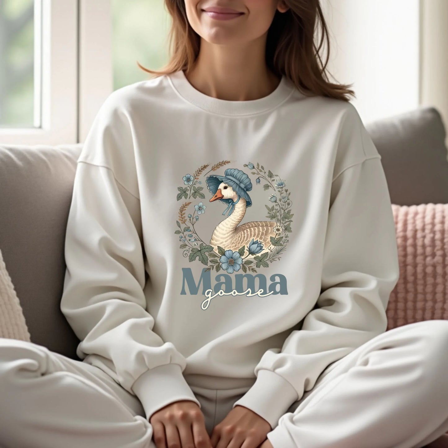 Woman sitting indoors wearing a white Mama Goose floral crewneck sweatshirt, highlighting the soft botanical goose illustration in a cozy setting.