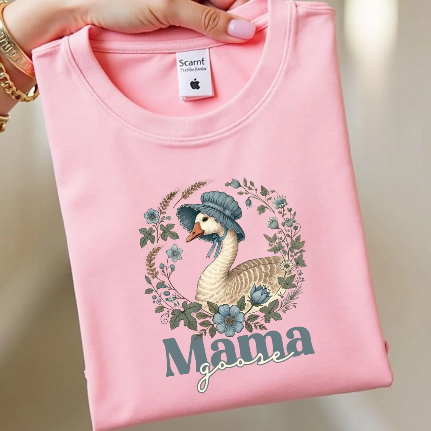 Flat lay of a pink Mama Goose floral t-shirt folded neatly, highlighting the vintage goose illustration surrounded by soft blue and green florals.
