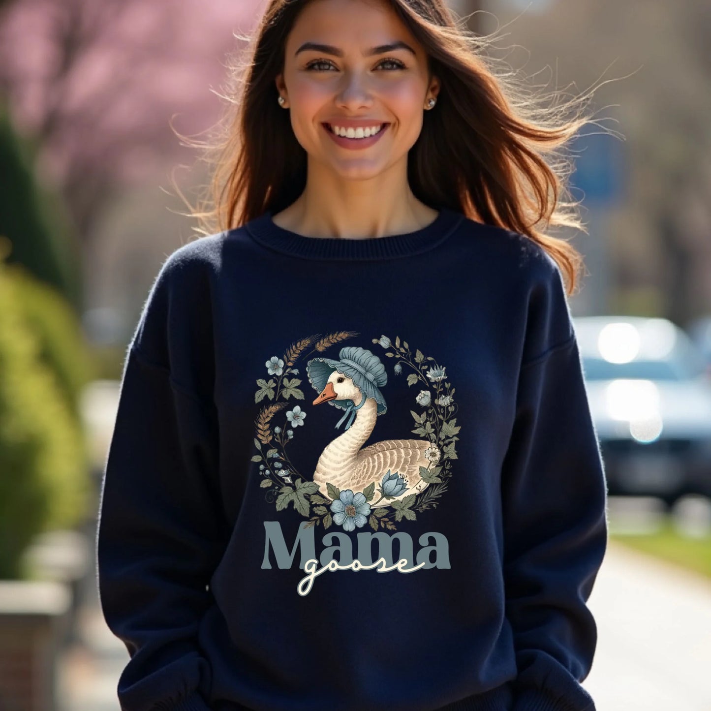 Smiling woman wearing a navy Mama Goose motherhood sweatshirt outdoors, featuring a floral wreath goose illustration with a cozy aesthetic.