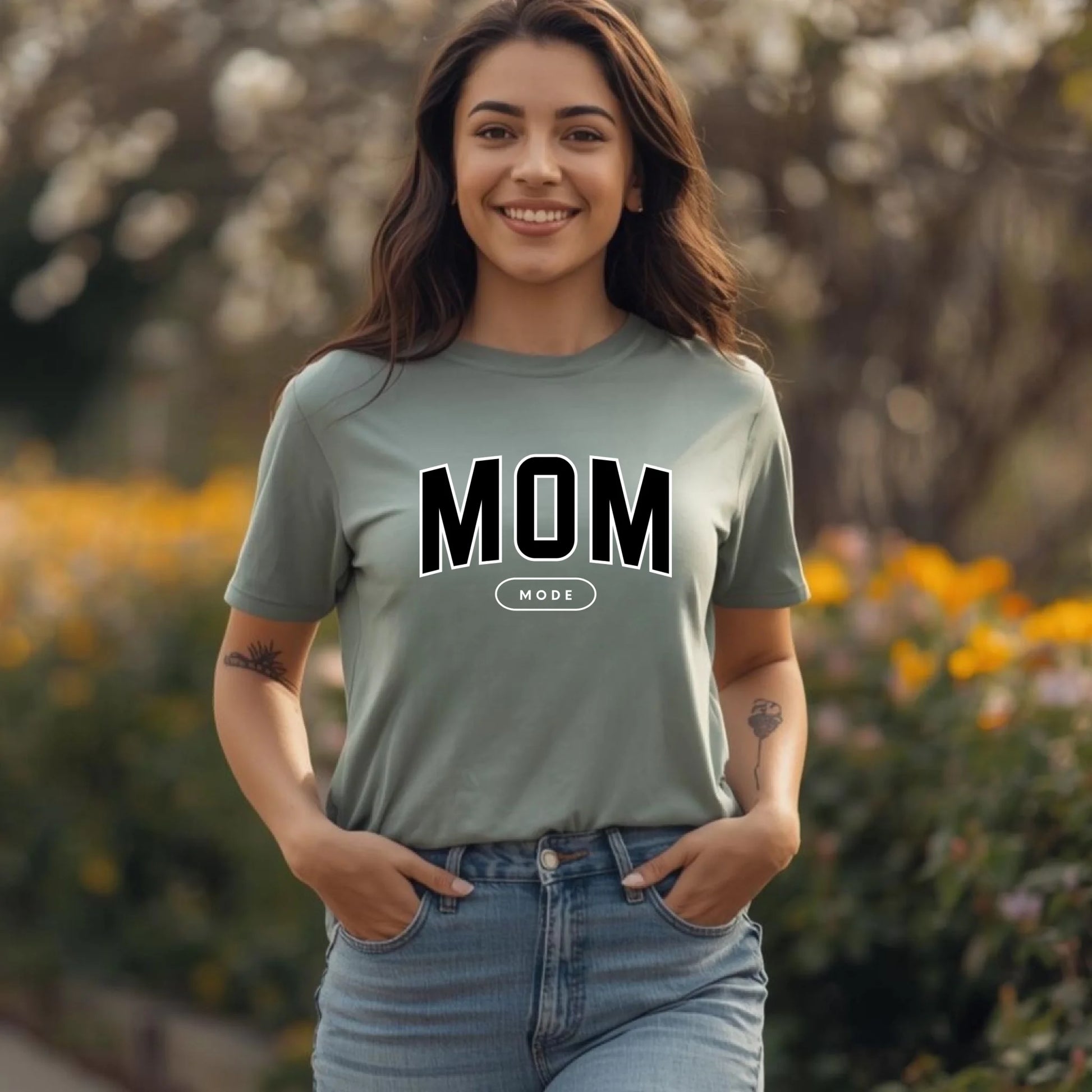 Woman wearing a 'MOM' t-shirt outdoors with blurred flowers and trees in the background