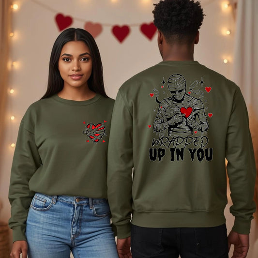 A couple wearing matching military green crewneck sweaters with a bold mummy design and Wrapped Up In You lettering on the back, romantic Valentine apparel.