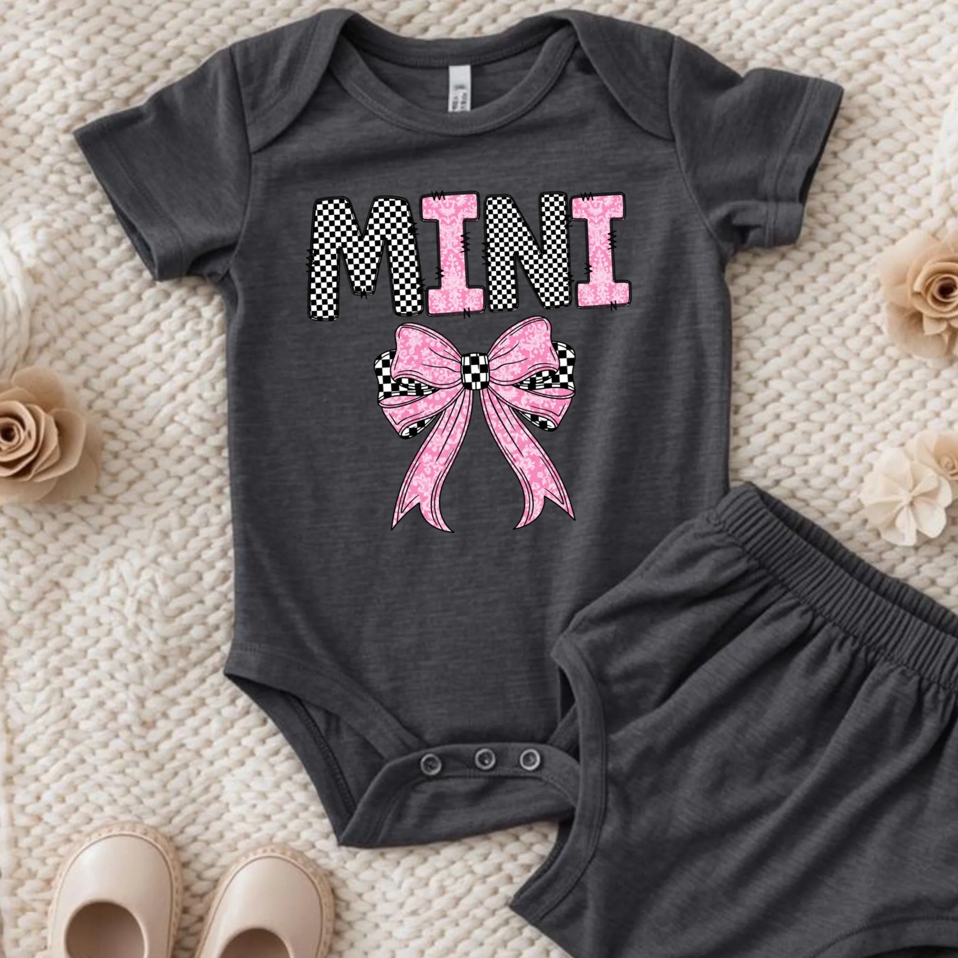 Dark gray baby onesie with pink bow and 'MINI' text on a textured surface.