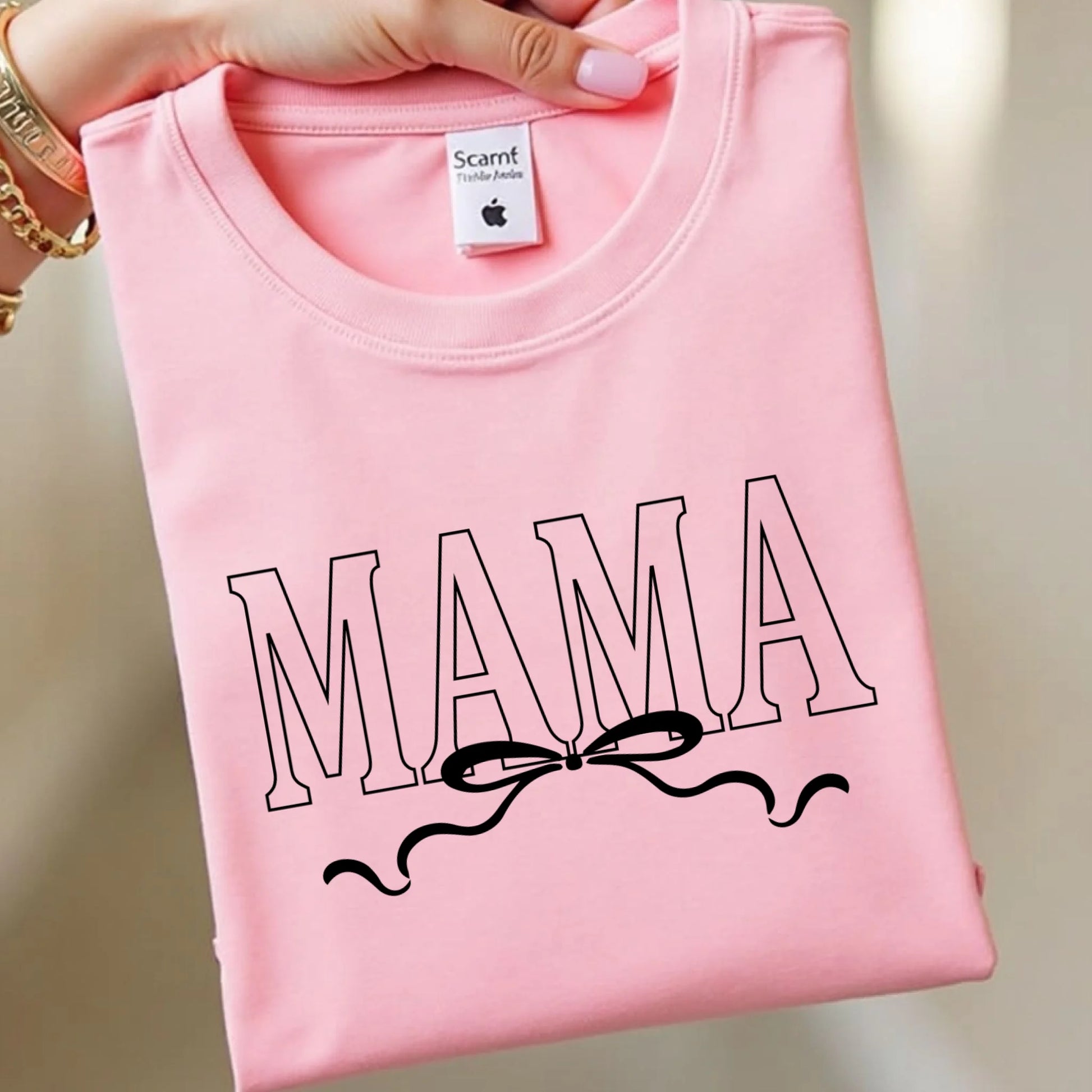 Pink t-shirt with 'MAMA' text and bow design, held by a hand.