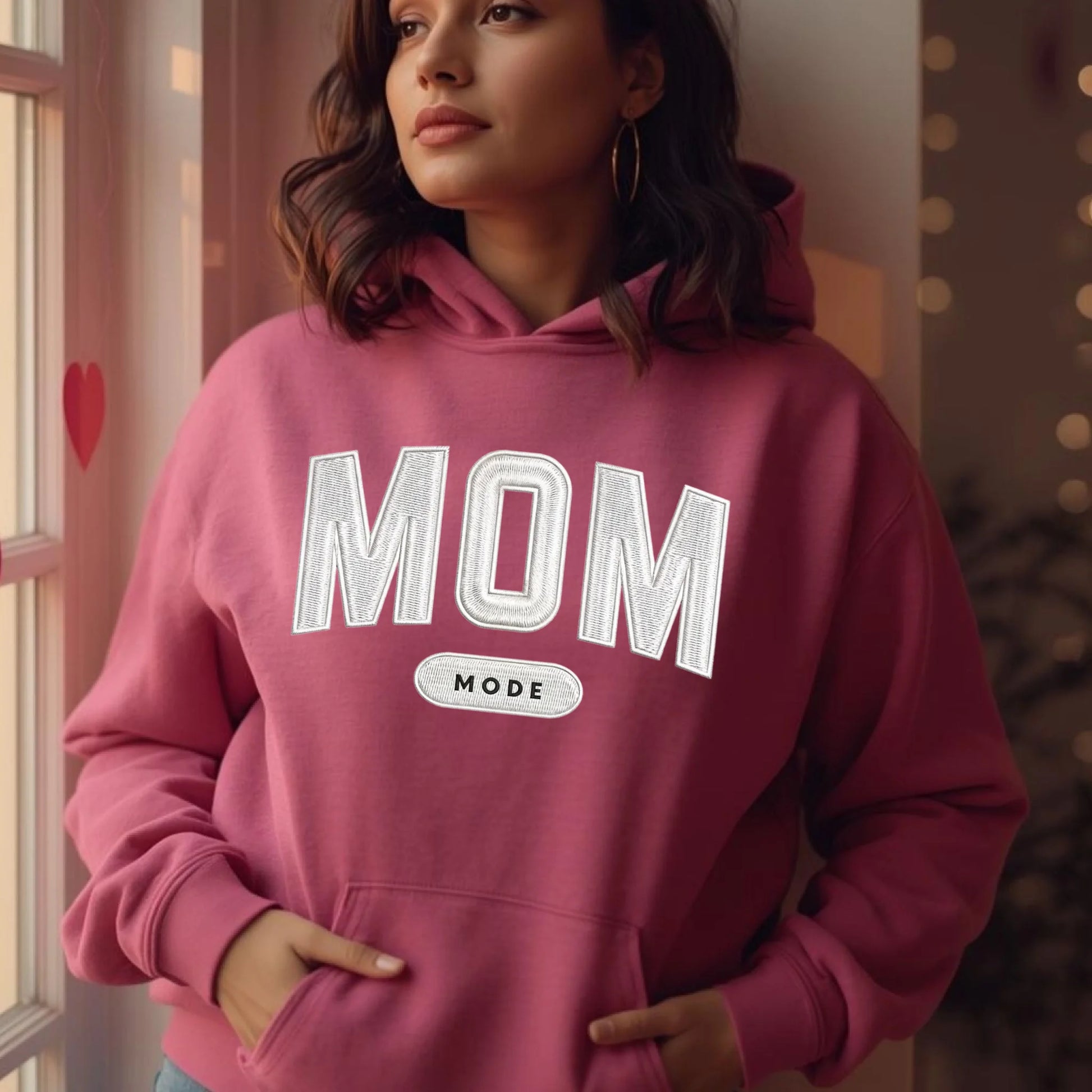 Woman wearing a azelea pink hoodie with 'MOM MODE' embroidered text, standing indoors.