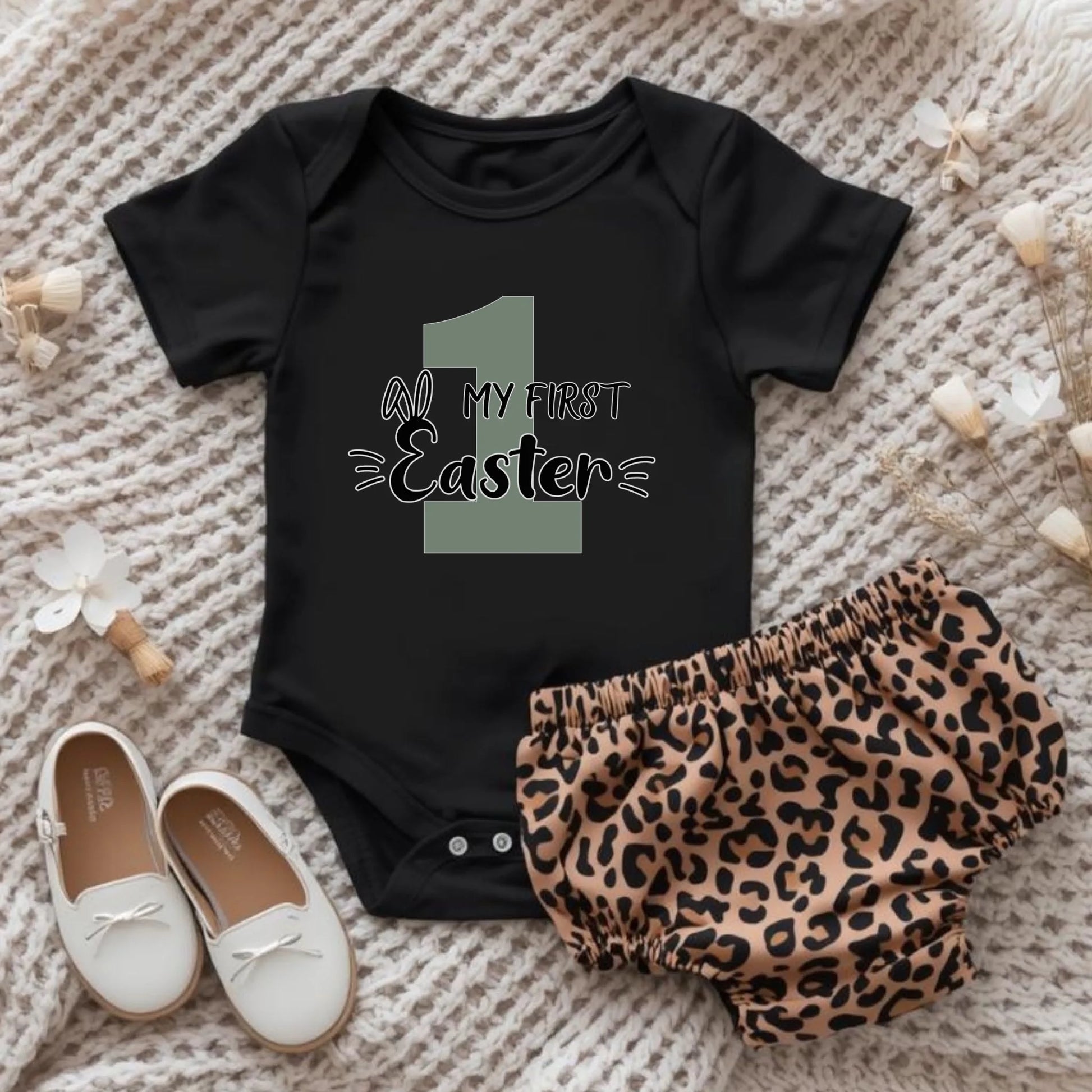 Flat lay of “My First Easter” infant onesie styled with neutral baby accessories, showcasing fit and design details.