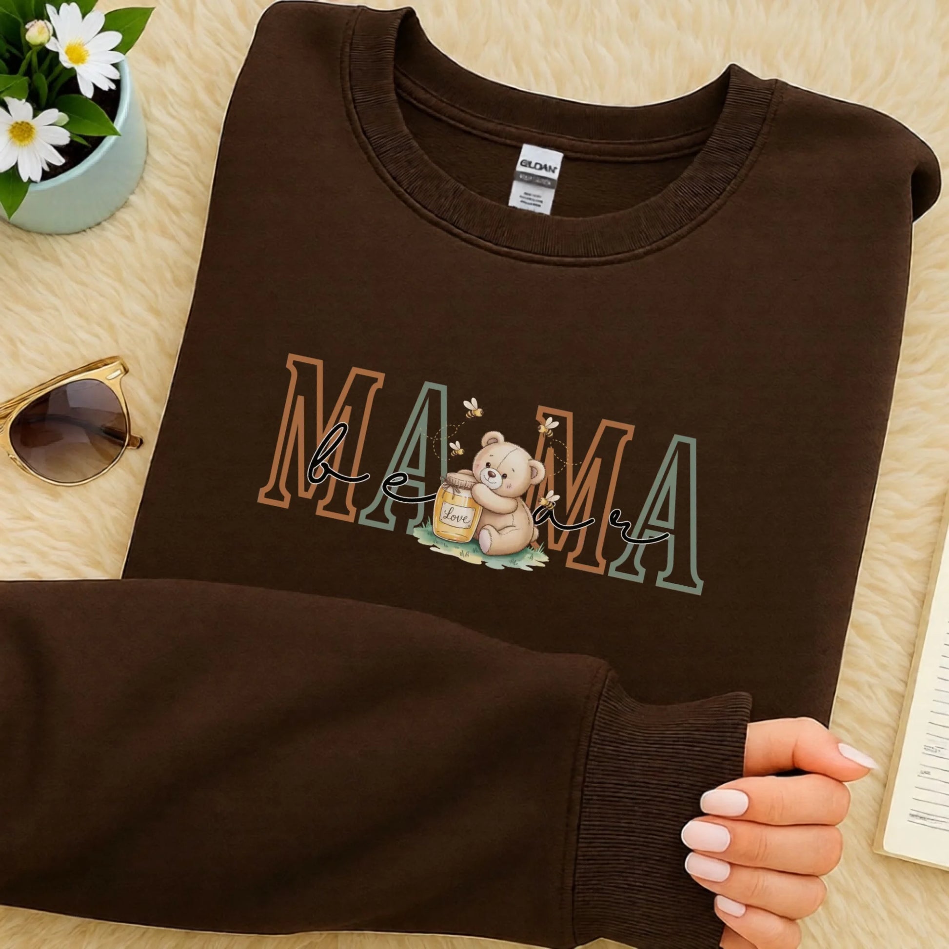 Folded Gildan crewneck sweatshirt featuring the Mama family role design with a teddy bear and honey illustration, shown in a rich brown colorway.