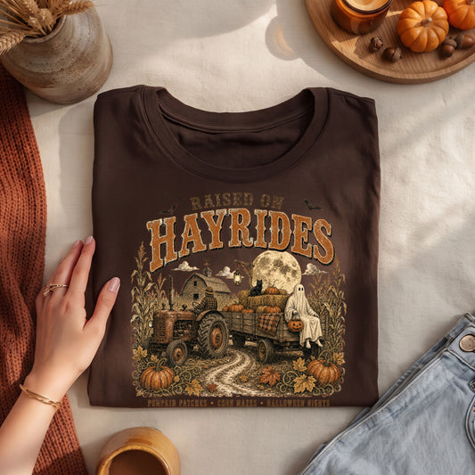 A folded dark brown graphic T-shirt is styled in a flat lay with a hand resting on the left side. The shirt features a detailed autumn harvest graphic with the text 'RAISED ON HAYRIDES' in large tan letters, showing a tractor, hayride wagon, ghosts, pumpkins, and a full moon.