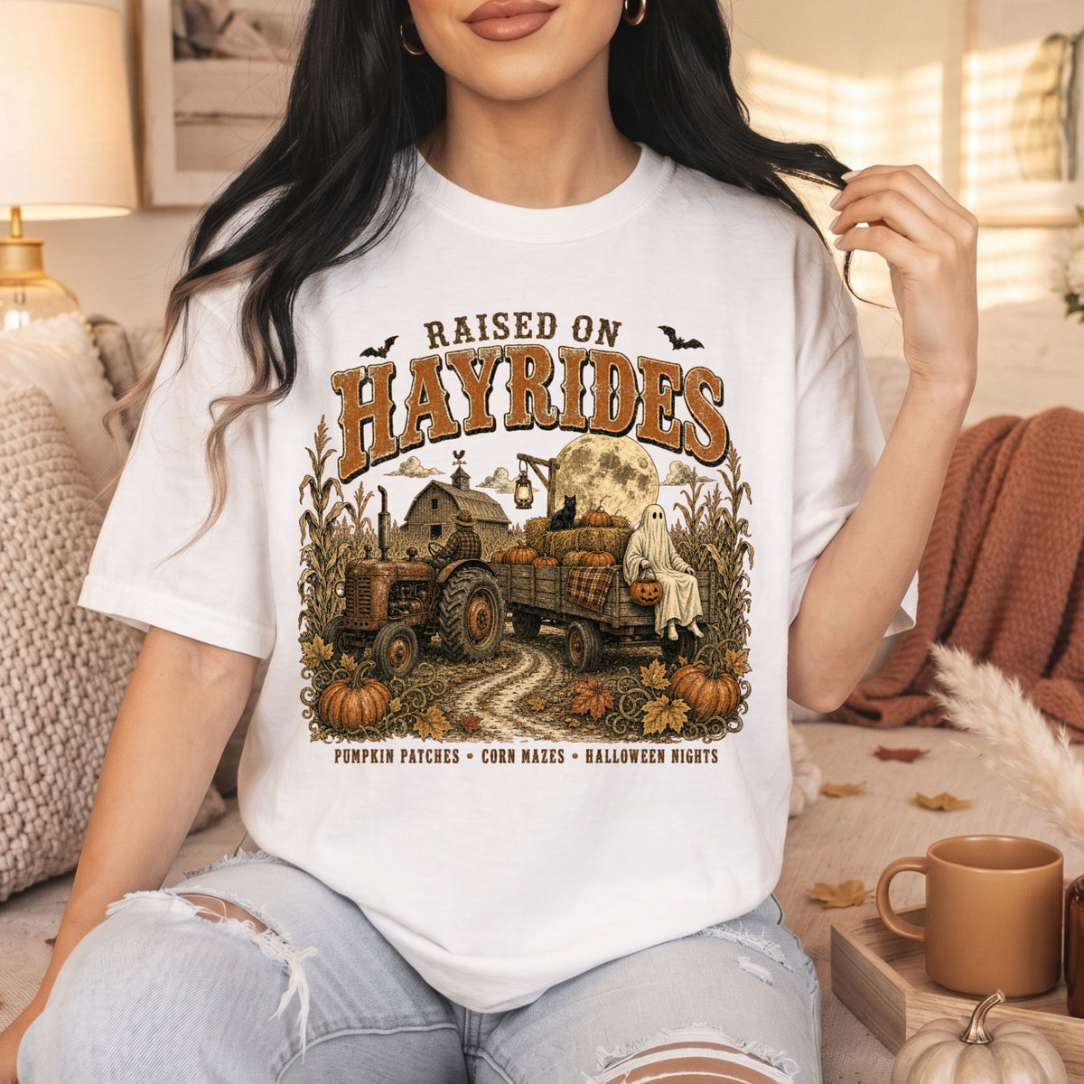 A woman wearing a white graphic t-shirt with a fall-themed 'Raised on Hayrides' design, sitting in a cozy living room setting.