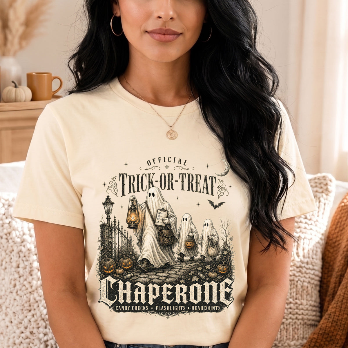 A woman wearing a cream-colored graphic t-shirt with a vintage-style Halloween design featuring ghosts, pumpkins, and the text 'Official Trick or Treat Chaperone'.