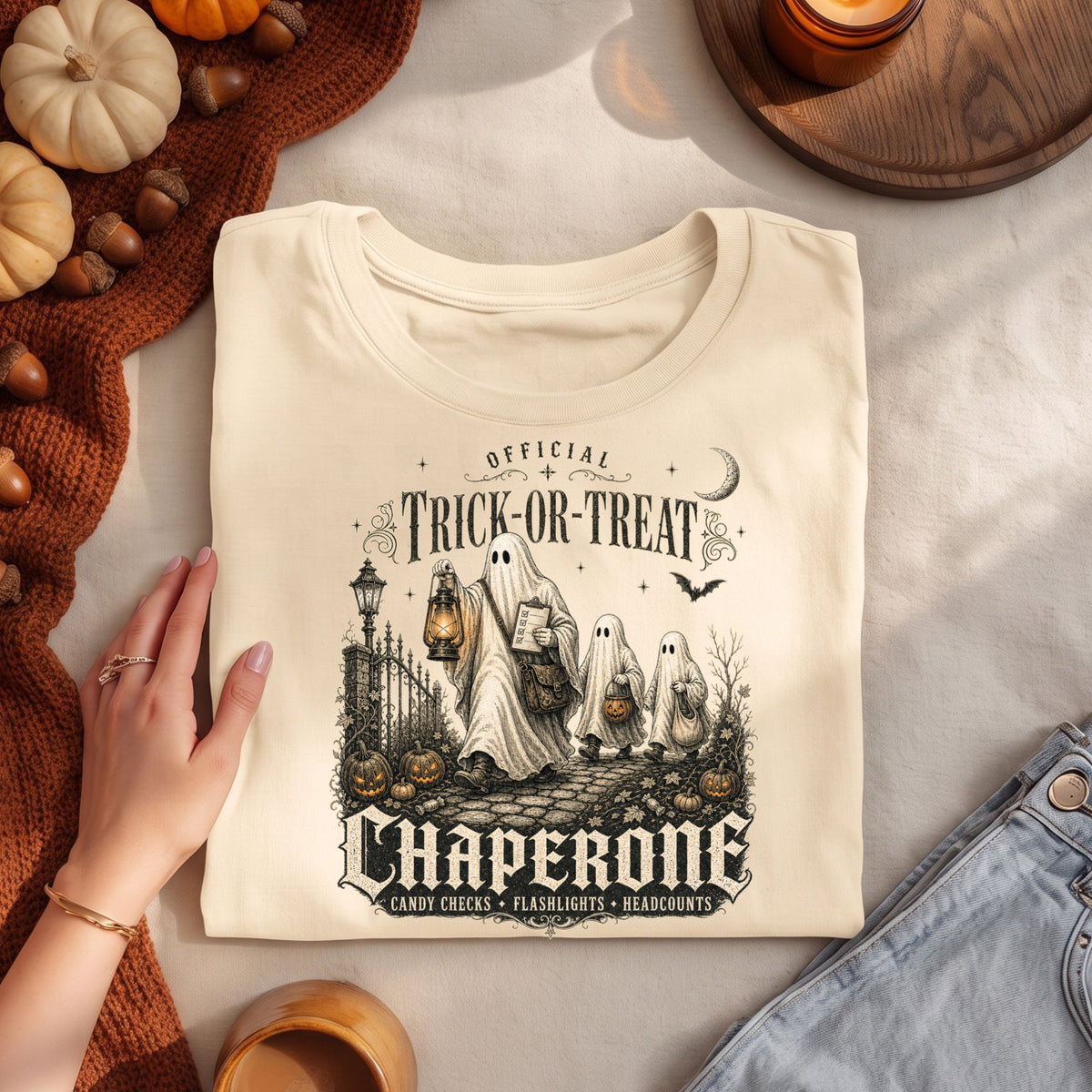 A folded cream-colored t-shirt with a Halloween-themed graphic print reading 'Official Trick or Treat Chaperone' is displayed on a light fabric surface. A hand is resting on the left side of the shirt, and autumn-themed props including pumpkins, a knit blanket, a candle, and denim jeans are arranged around it.