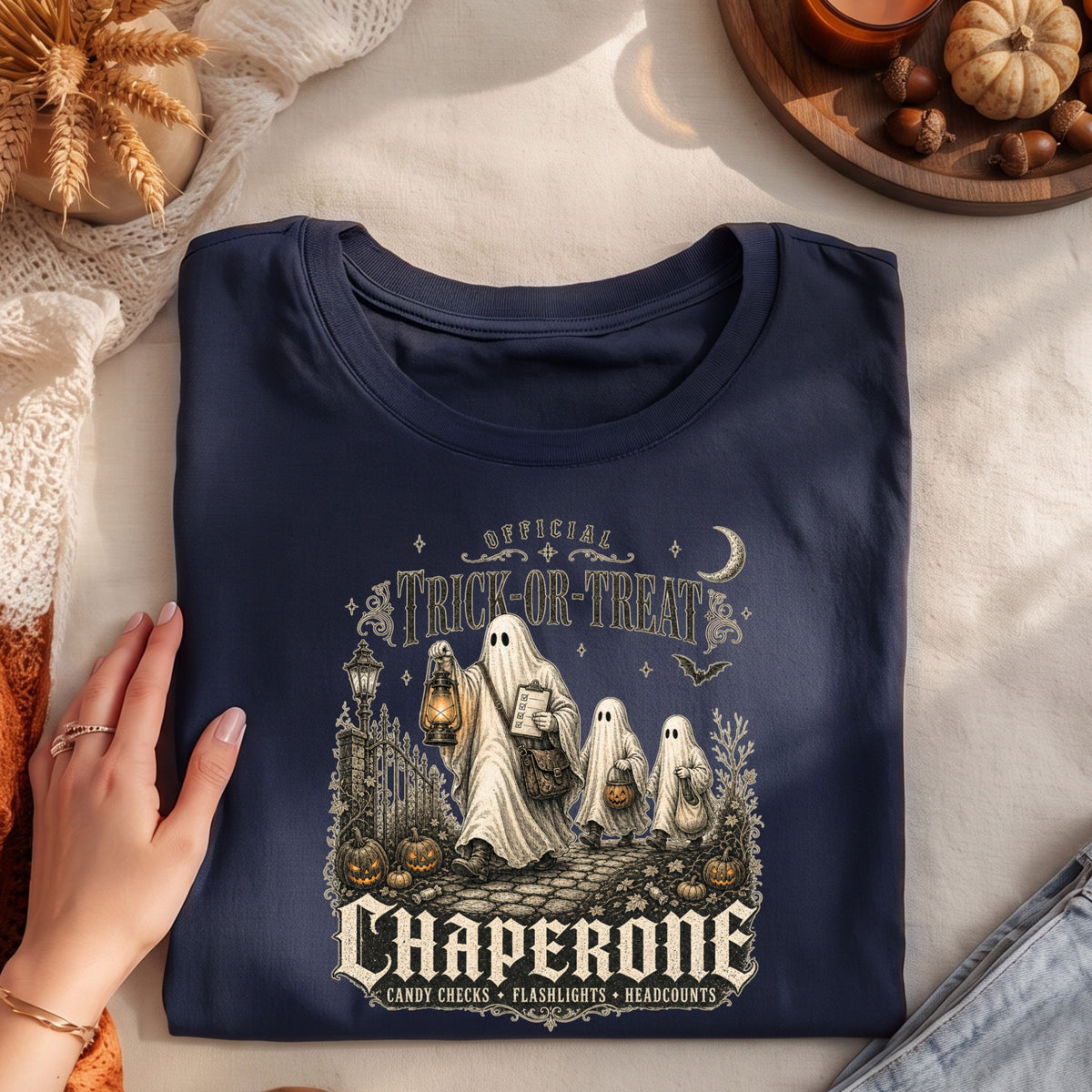 A navy blue folded graphic T-shirt is displayed on a light fabric surface with a hand resting on the left side. The shirt features a Halloween-themed illustration with three ghosts, pumpkins, lanterns, a crescent moon, and the text 'Official Trick or Treat Chaperone' and 'Candy Checks • Flashlights • Beacons.