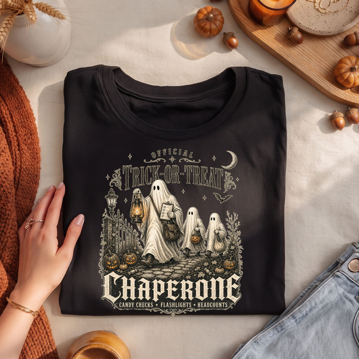 A folded black graphic T-shirt is displayed in a styled flat lay with a hand resting on the left side. The shirt features a vintage-style Halloween illustration with ghosts, pumpkins, and the text 'Official Trick or Treat Chaperone'.