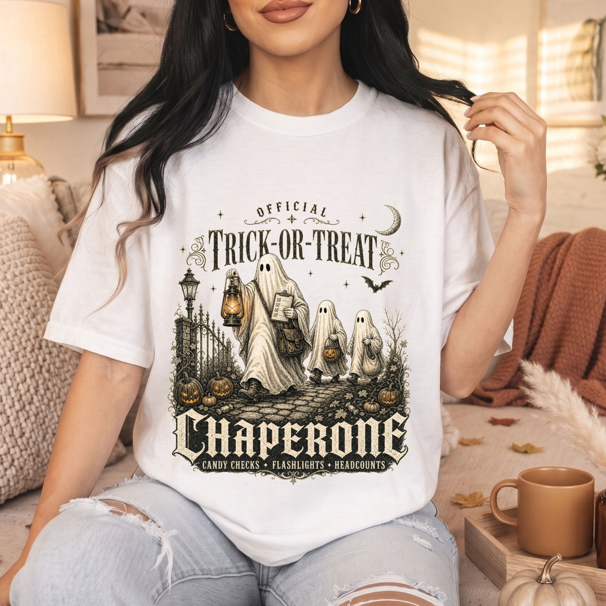 A woman wearing a white graphic t-shirt with a Halloween-themed design that reads 'Official Trick or Treat Chaperone'. The scene is set in a cozy, warm-toned living room with autumn decorations, a mug, and a lamp.