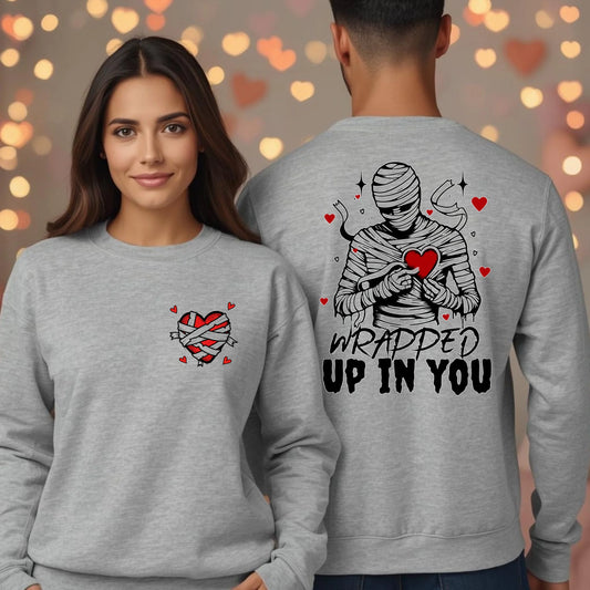 A woman wearing a grey crewneck with a wrapped heart front graphic stands next to a man showing the mummy back design with the words Wrapped Up In You, Valentine sweatshirt.