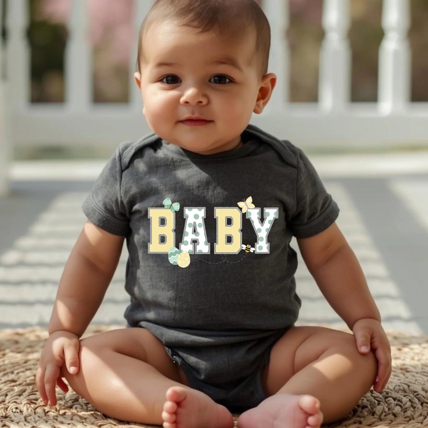 Baby wearing a dark gray “Baby” Easter t-shirt from a Mommy and Me matching family set, seated outdoors.