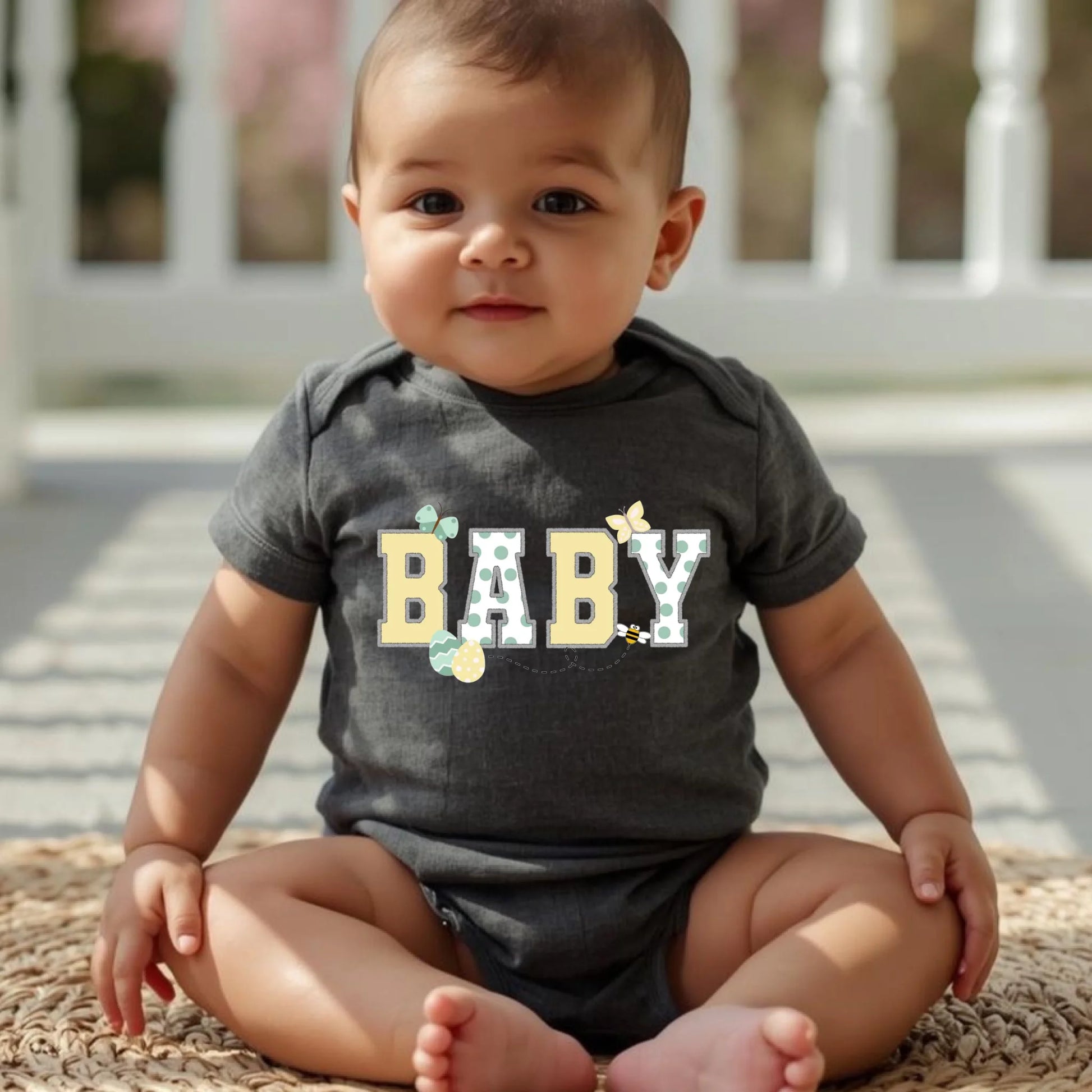 Baby wearing a dark gray “Baby” Easter t-shirt from a Mommy and Me matching family set, seated outdoors.