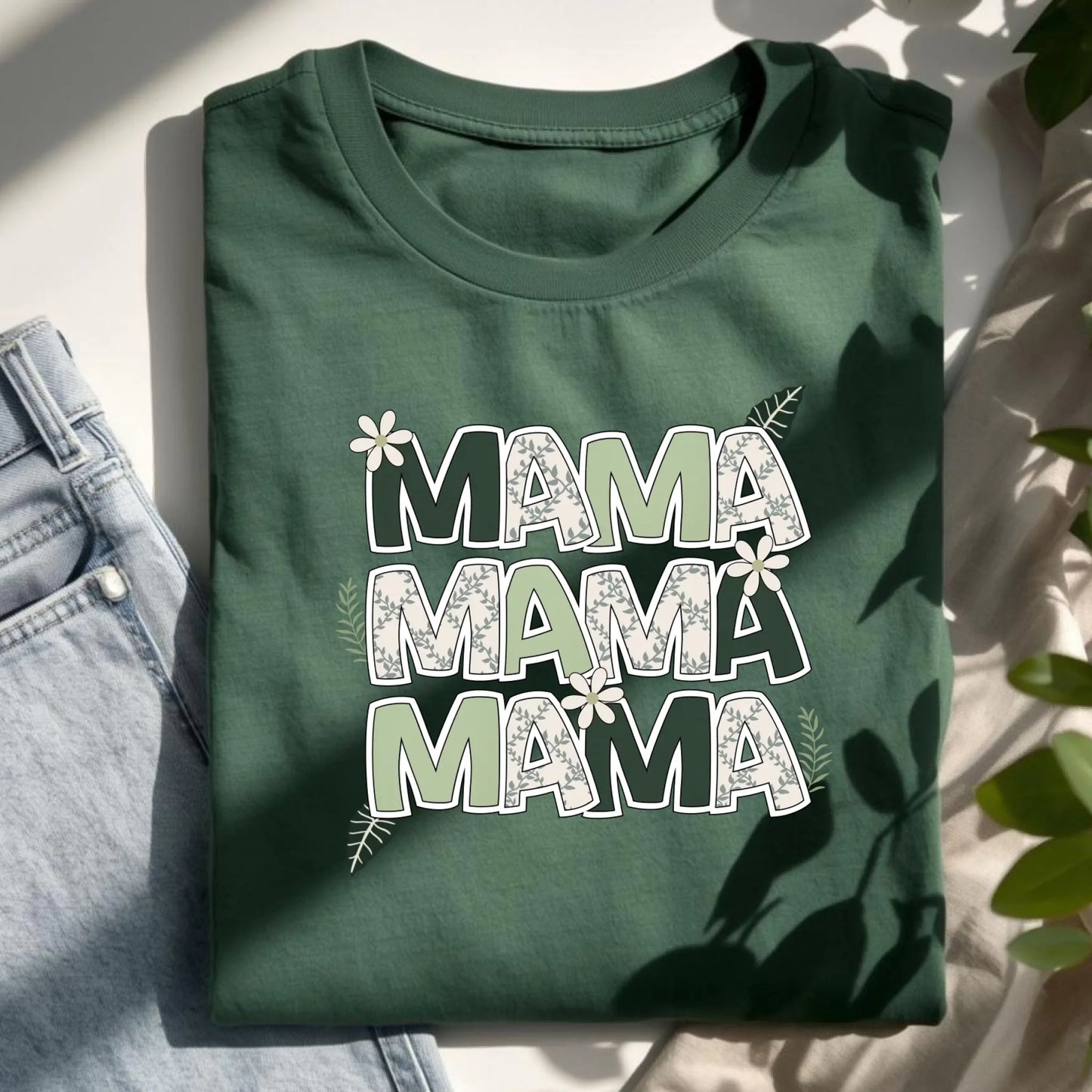 Flat lay of a green Bella Canvas t-shirt with a floral “Mama” design styled with denim and neutral accents.