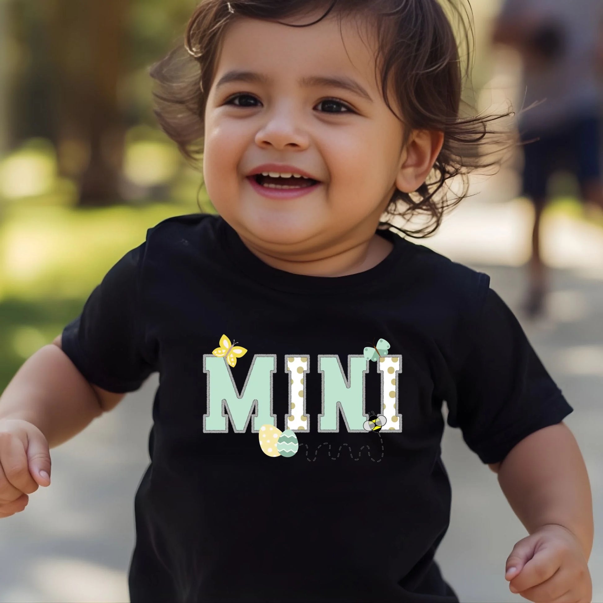 Toddler wearing a black “Mini” Easter t-shirt from a Mommy and Me matching collection, smiling outdoors.