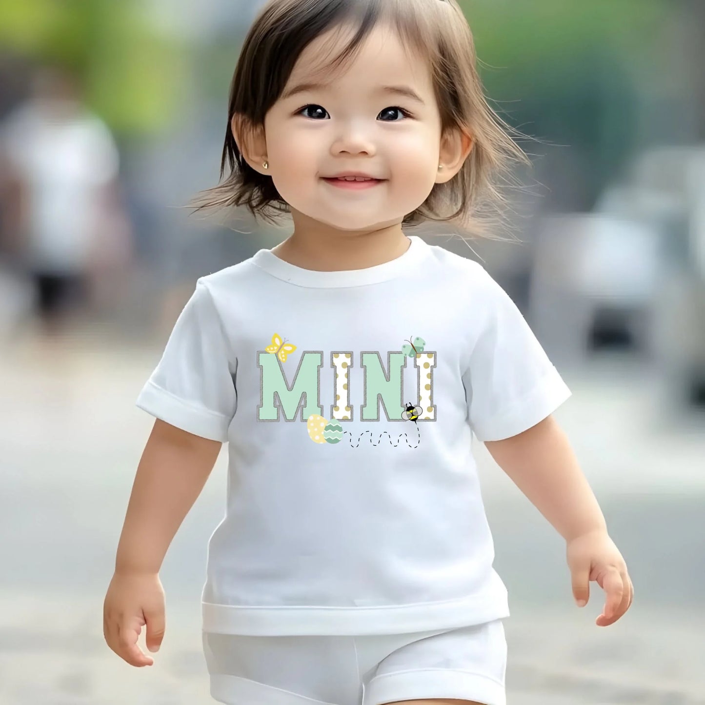 Toddler wearing a white “Mini” Easter t-shirt from a Mommy and Me matching set, walking outdoors in a spring lifestyle photo.