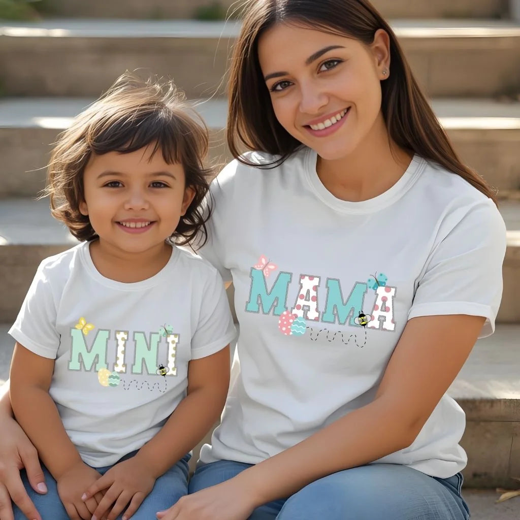 Mother and young child wearing matching Mommy and Me Easter t-shirts with “Mama” and “Mini” designs in soft spring colors, seated together outdoors.
