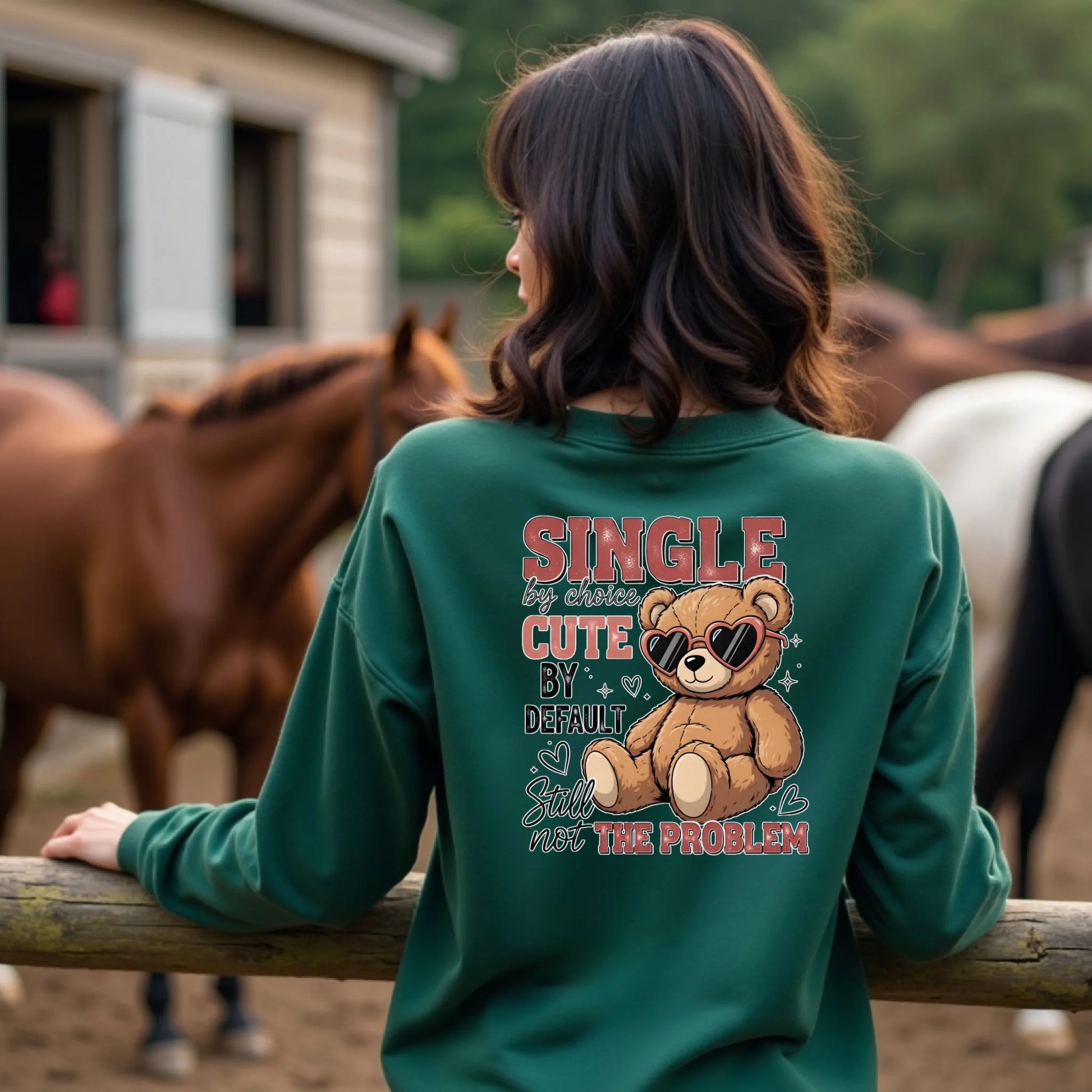 Person wearing a green sweatshirt with a teddy bear graphic and text, standing in front of horses.