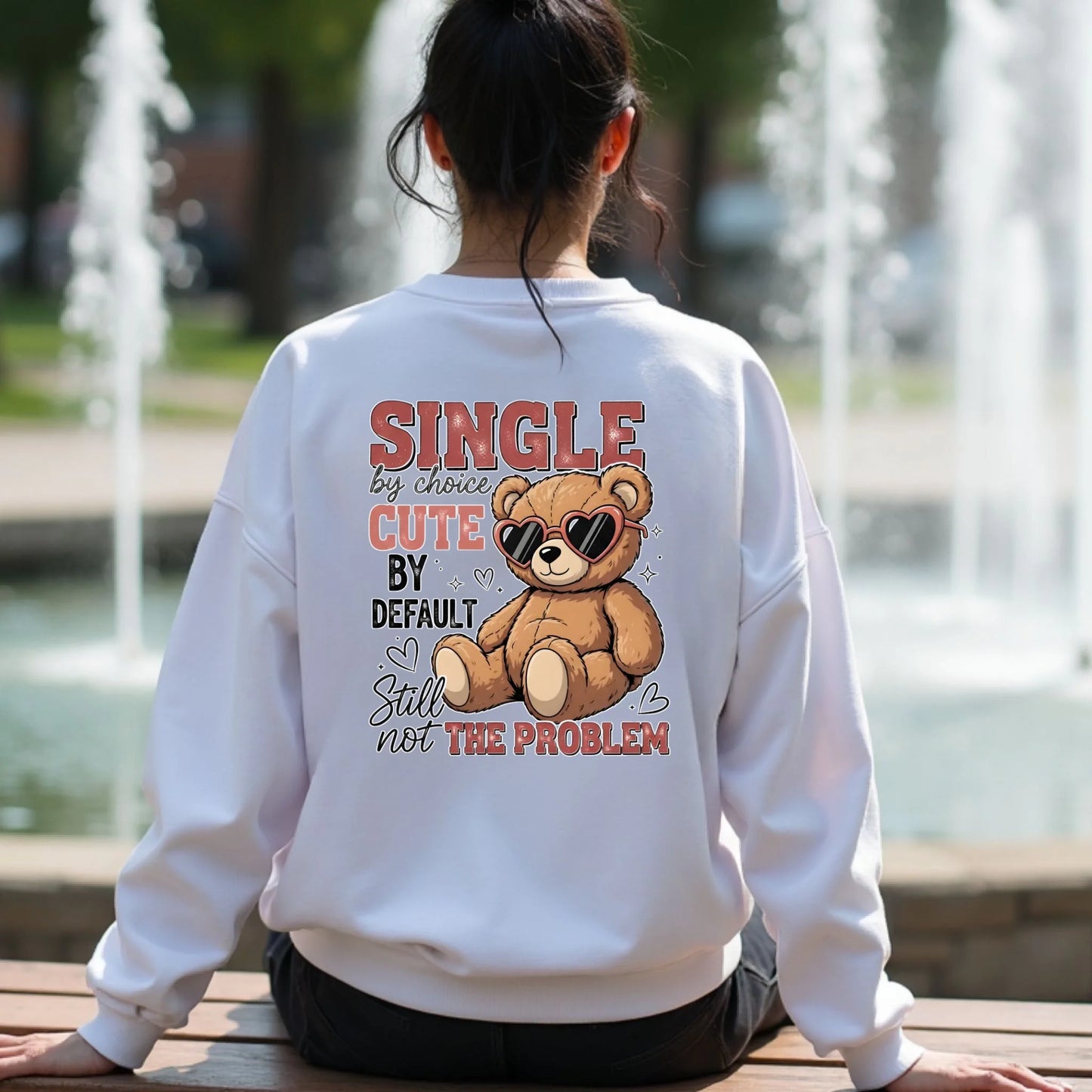 Person wearing a white sweatshirt with a teddy bear graphic and text by a fountain.
