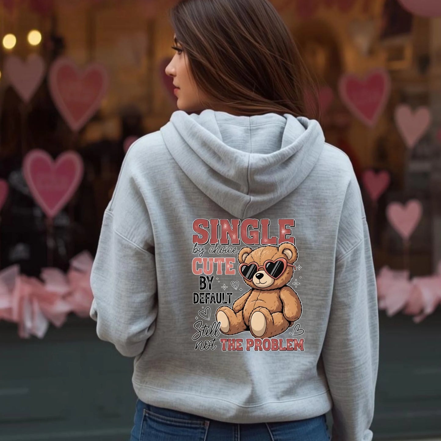 Person wearing a gray hoodie with a teddy bear graphic and text in an urban setting with heart decorations.