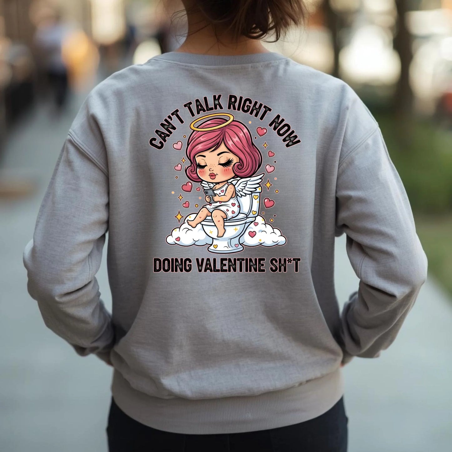 Person wearing a sport gray crewneck sweatshirt with a graphic design on the back, featuring a humorous Toilet Cupid Valentine graphic.