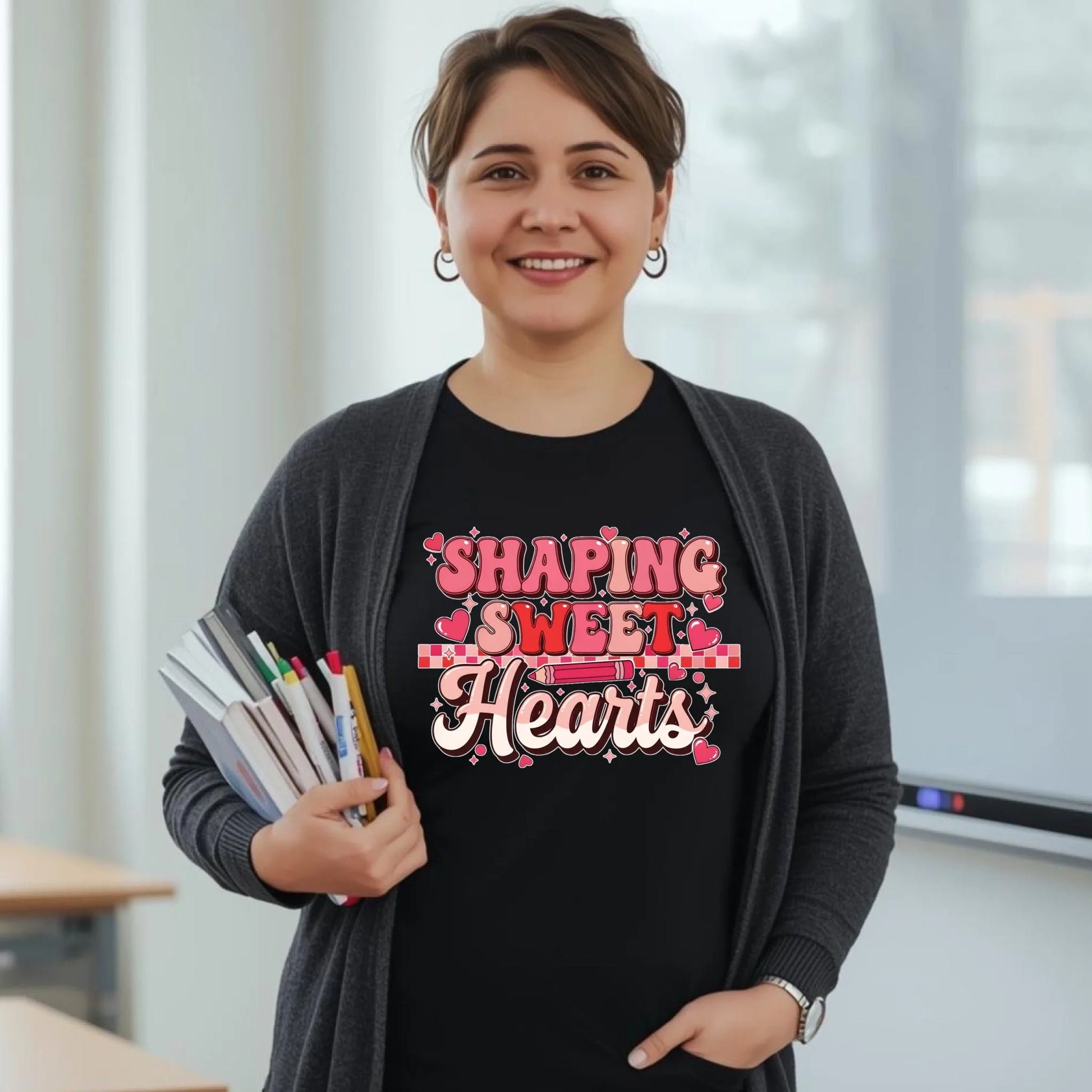 Teacher wearing a black “Shaping Sweet Hearts” shirt and holding colorful markers in a classroom. Cute Valentine school outfit.